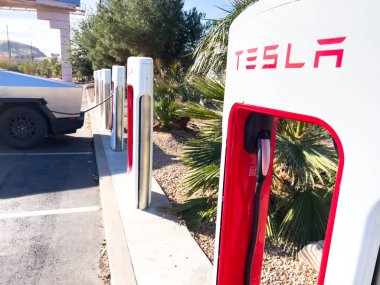 Mesquite, Nevada, ABD-10 Kasım 2024 - Tesla Cybertruck 'ın bir Tesla Superşarj istasyonuna bağlı yan görüntüsü, açık mavi gökyüzü altında aracın parlak tasarımını gösteriyor. Palmiye ağaçları ve yakınlarda