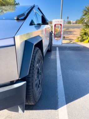 Mesquite, Nevada, ABD-10 Kasım 2024 - Tesla Cybertruck 'ın bir Tesla Superşarj istasyonuna bağlı yan görüntüsü, açık mavi gökyüzü altında aracın parlak tasarımını gösteriyor. Palmiye ağaçları ve yakınlarda