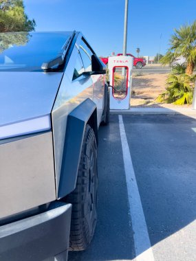 Mesquite, Nevada, ABD-10 Kasım 2024 - Tesla Cybertruck 'ın bir Tesla Superşarj istasyonuna bağlı yan görüntüsü, açık mavi gökyüzü altında aracın parlak tasarımını gösteriyor. Palmiye ağaçları ve yakınlarda