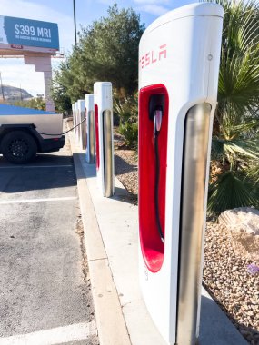 Mesquite, Nevada, ABD-10 Kasım 2024 - Tesla Cybertruck 'ın bir Tesla Superşarj istasyonuna bağlı yan görüntüsü, açık mavi gökyüzü altında aracın parlak tasarımını gösteriyor. Palmiye ağaçları ve yakınlarda