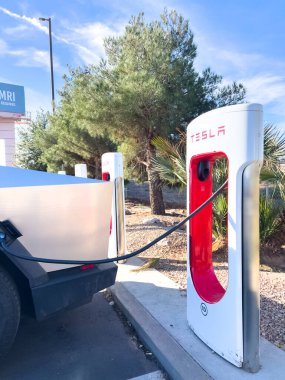 Mesquite, Nevada, ABD-10 Kasım 2024 - Tesla Cybertruck 'ın bir Tesla Superşarj istasyonuna bağlı yan görüntüsü, açık mavi gökyüzü altında aracın parlak tasarımını gösteriyor. Palmiye ağaçları ve yakınlarda