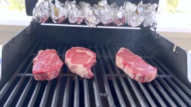 Bu resim barbekü ızgarasında pişen üç kalın bifteği, yukarıdaki koçanda folyoya sarılı mısırla birlikte, tipik bir açık hava yemek sahnesini görüntülüyor.