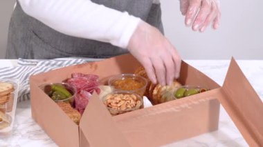 Elleri titizlikle bir kaseye taze kırmızı üzüm ekliyor, çeşitli peynirler, zeytinler ve pişirilmiş etlerle dolu güzel bir şarküteri kutusunu tamamlıyor.
