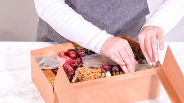 Elleri titizlikle bir kaseye taze kırmızı üzüm ekliyor, çeşitli peynirler, zeytinler ve pişirilmiş etlerle dolu güzel bir şarküteri kutusunu tamamlıyor.