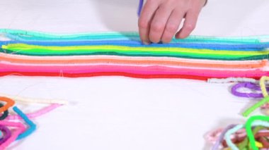 Eller birbirine zıt beyaz bir yüzeye karşı düzgün düzenlenmiş renkli, sarmalanmış kil boncuk iplikçiklerine dokunur..