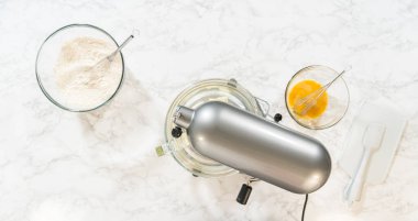 Top view of an electric mixer and bowls containing ingredients for Classic Sugar Cookie Dough. The mixer is placed over a bowl with partially mixed wet ingredients, while nearby bowls hold flour and a clipart