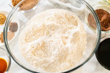 A large glass mixing bowl filled with sifted flour, placed on a marble surface along with other ingredients like spices, butter, and molasses for the Chocolate Gingerbread Cookie Dough. clipart