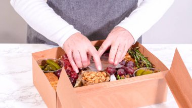 Elleri titizlikle bir kaseye taze kırmızı üzüm ekliyor, çeşitli peynirler, zeytinler ve pişirilmiş etlerle dolu güzel bir şarküteri kutusunu tamamlıyor.