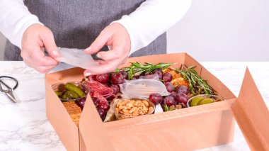 Elleri titizlikle bir kaseye taze kırmızı üzüm ekliyor, çeşitli peynirler, zeytinler ve pişirilmiş etlerle dolu güzel bir şarküteri kutusunu tamamlıyor.