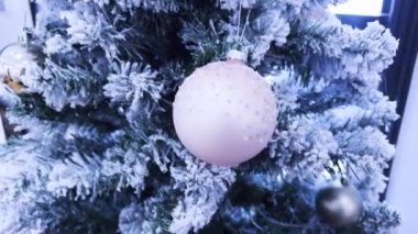 Ön planda, ışıltılı süslemelerle süslenmiş karla kaplı bir Noel ağacı duruyor. Arkasında, şık bir yaşam alanı beyaz bir kanepe, yeşil bir duvar ve neşeli bir mevsimlik dekor barındırır.