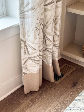 Hassas bir çizgili deseni olan beyaz perdelerin yakın çekimi, uzaya doku ve zarafet katıyor. Perdeler bir pencerenin yakınındaki ahşap zemine nazikçe örtülmüş.