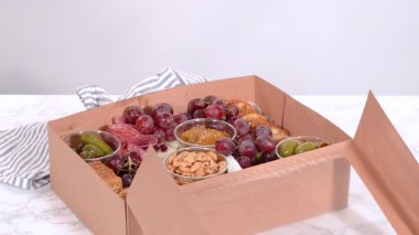 Elleri titizlikle bir kaseye taze kırmızı üzüm ekliyor, çeşitli peynirler, zeytinler ve pişirilmiş etlerle dolu güzel bir şarküteri kutusunu tamamlıyor.