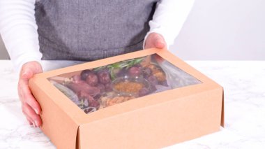 Elleri titizlikle bir kaseye taze kırmızı üzüm ekliyor, çeşitli peynirler, zeytinler ve pişirilmiş etlerle dolu güzel bir şarküteri kutusunu tamamlıyor.