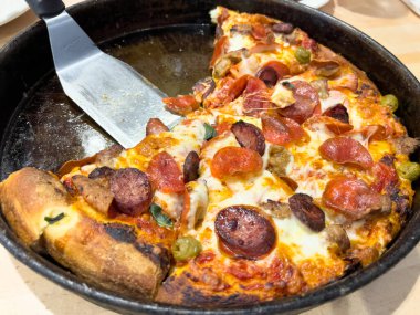 Eritilmiş peynir, pepperoni, sosis, yeşil zeytin ve fesleğenle kaplanmış dökme demir bir tavada yeni pişmiş kalın tabak pizza. Kıtır kıtır ve yapışkan peynir bunu ağız sulandırıcı yapar.