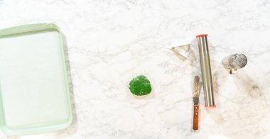 Overhead view of Green Gingerbread Cookies dough, partially rolled out, next to tree-shaped cutouts on a baking sheet and baking tools. clipart