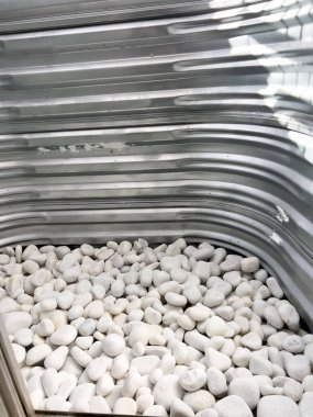 Altında beyaz dekoratif taşlar olan kıvrımlı metal bir pencerenin yakından görüntüsü. Metal yüzeyde doğal ışığı yansıtan küçük çizikler ve aşınmalar var. Bu işlevsel tasarım genellikle