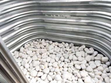 Altında beyaz dekoratif taşlar olan kıvrımlı metal bir pencerenin yakından görüntüsü. Metal yüzeyde doğal ışığı yansıtan küçük çizikler ve aşınmalar var. Bu işlevsel tasarım genellikle
