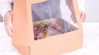 Elleri titizlikle bir kaseye taze kırmızı üzüm ekliyor, çeşitli peynirler, zeytinler ve pişirilmiş etlerle dolu güzel bir şarküteri kutusunu tamamlıyor.