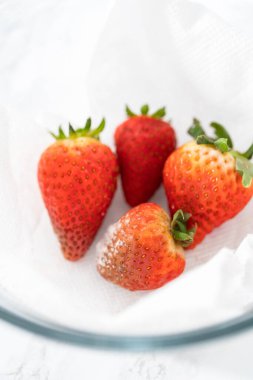 Parlak kırmızı çilekler, küf izleriyle karışmış, beyaz peçetedeki kağıt havluyla kaplı cam bir kasede dinleniyor, depolama tekniklerini gösteriyor..