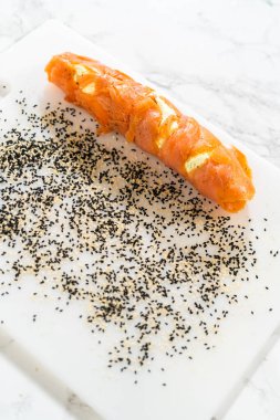 Tütsülenmiş somon rulosu siyah ve beyaz susam tohumlarının karışımının üzerine yuvarlanarak lezzetli ve dokulu bir kaplama oluşturur..