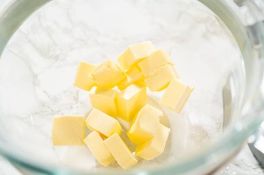 Küçük tuzsuz tereyağı küpleri cam bir karıştırma kabına, mermer bir tezgahın üzerine yerleştirilmiş. Bu malzeme hazırlık aşısı yemek tarifleri, yemek pişirme dersleri ve yemek tarifleri için idealdir.