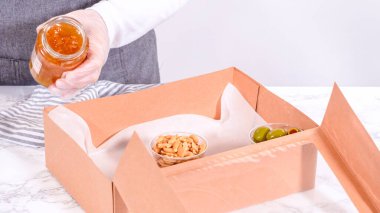 Elleri titizlikle bir kaseye taze kırmızı üzüm ekliyor, çeşitli peynirler, zeytinler ve pişirilmiş etlerle dolu güzel bir şarküteri kutusunu tamamlıyor.