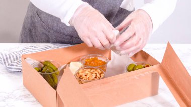 Elleri titizlikle bir kaseye taze kırmızı üzüm ekliyor, çeşitli peynirler, zeytinler ve pişirilmiş etlerle dolu güzel bir şarküteri kutusunu tamamlıyor.