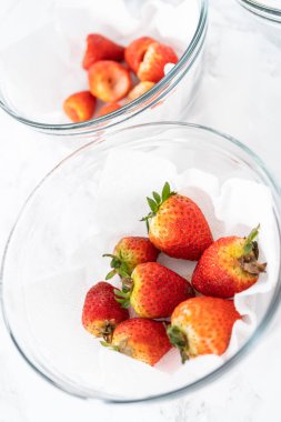 Parlak kırmızı çilekler, küf izleriyle karışmış, beyaz peçetedeki kağıt havluyla kaplı cam bir kasede dinleniyor, depolama tekniklerini gösteriyor..