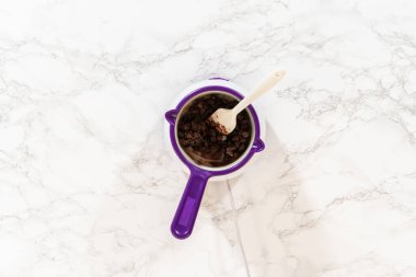 Beyaz mermer bir yüzeye kurulmuş çikolata parçacıkları ve bir parça tereyağı ile dolu mor bir eritme kabının genel görünümü. Bu adım Easy Last Minute Ren Geyiği Şekerli Kurabiye 'nin yapımının bir parçası.