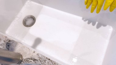 Sarı eldivenli ellerin bulaşıkları yıkadığı, köpüklerle çevrili bir lavaboda özenle ovaladığı, temizlik aletleri ve temiz su bardakları ile görülmektedir..