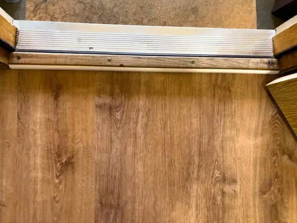 Ahşap döşeme ve beton yüzey arasındaki geçişi gösteren iç kapı eşiğini yakından görüyor. Metal şerit ve çevreleyen çerçeve, doku ve inşaat detaylarını vurgular