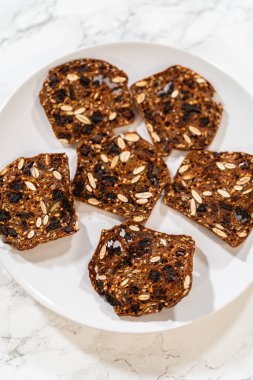 Beyaz bir tabakta tohum ve kurutulmuş meyve içeren çok tahıllı krakerlerin yakın çekim görüntüsü. Bakış açısı zengin dokuları ve çıtır çıtır kenarları vurgular, onları peynir için mükemmel yapar.