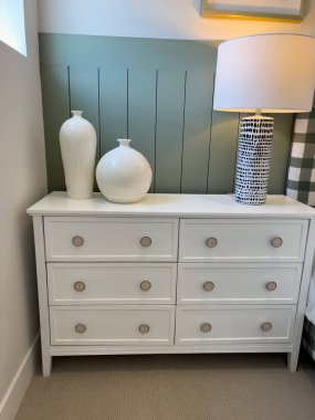 Şık bir yatak odası köşesinde minimalist ahşap aksanlı düğmeleri olan altı çekmeceli beyaz bir şifonyer. Şifonyerin üzerinde iki modern seramik vazo ve adaçayı yeşili bir masa lambası var.