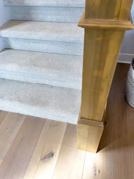 Hafif halı döşenmiş bir merdivenin yakın çekimi doğal ahşap yeni bir direkle tahta döşemeye dönüşüyor. Görüntü modern bir evdeki döşeme malzemeleri arasındaki temiz kavşağı vurgular.