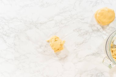 Klasik Şekerli Kurabiye Hamuru 'nun mermer bir yüzeyde plastik gıda ambalajına sarılı halinin en üst görüntüsü. Daha büyük bir hamur topu buzdolabında soğutulmaya hazırdır. Hamuru sağlamlaştırmak için gerekli bir adımdır.