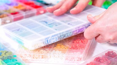 Kadın elleri şeffaf organizatörlerin renklerine göre sıralanmış boncuk koleksiyonunun üzerinde zarif bir şekilde duruyor. Boncuk dizisi, derin morlardan parlak portakallara kadar, titizlikle geniş bir yelpazeyi kapsıyor.