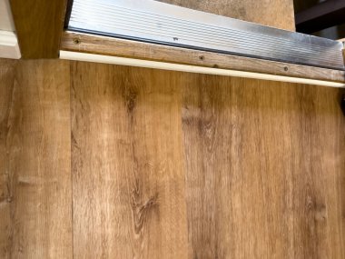 Close-up view of a door threshold where a light brown wood floor meets a raised metal edge and a concrete step. The worn wooden transition strip and screw-mounted metal trim show subtle texture and