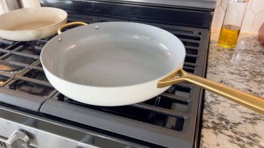 Siyah bir gaz sobasının üzerinde duran altın saplı beyaz, yapışmaz bir tavanın yakın çekimi. Arka planda ikinci bir tencere ve granit tezgahta bir şişe yemek yağı görülebilir. Sahne