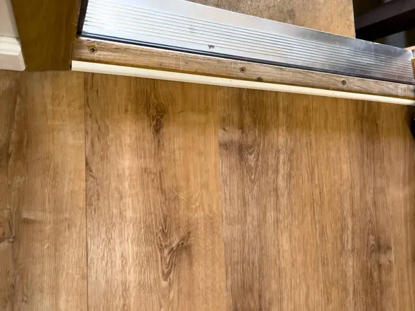 Açık kahverengi ahşap zeminin yükseltilmiş metal bir kenar ve beton bir basamakla buluştuğu kapı eşiğinin yakın görüntüsü. Yıpranmış ahşap geçiş şeridi ve vida monte edilmiş metal süslemesi ince bir doku ve...