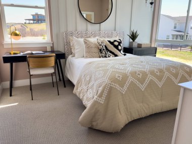 Parlak, modern bir yatak odası bej bir yorgan, yatak başlığının üstündeki ayna ve güneşli bir dış görünüş sunan büyük pencereler..