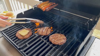 Birkaç hamburger köftesi ve sosis pişirilmiş bir gaz ızgarasının yakın çekimi. Üstünde bir dilim peynirin eridiği bir köfte de dahil. Metal maşa tutan bir el, hamburgeri görünür hale getiriyor.