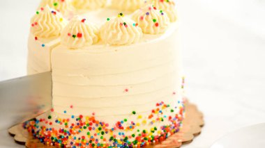 Bıçak, üzerine renkli süslemeler ve borulu krema serpiştirilmiş beyaz kremalı pastayı keser. Pasta, mermer mutfak tezgahının üzerinde altın bir tahtanın üzerinde duruyor..