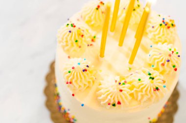 Bir doğum günü pastasının kıvrımlı şekerlemesine yerleştirilmiş birkaç sarı mumla açılı üst görüntüsü. Altta ve üstte renkli serpintiler var. Mumlardan hafif bir gölge görünüyor..