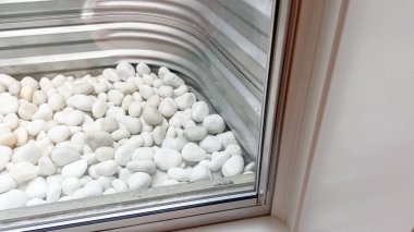 Beyaz dekoratif taşlar, içeriden iyi görünen alüminyum bir pencereyi dolduruyor. Temiz düzenleme ve parlak ışık minimalist ve modern bir izlenim veriyor..