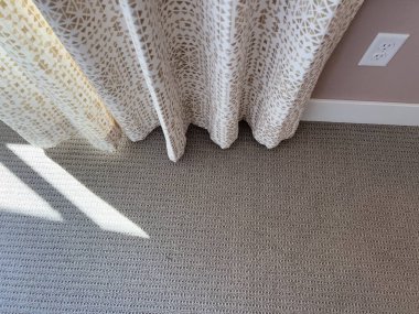 Modern bir odada yumuşak güneş ışığının dokulu nötr bir halının üzerine düşmesine izin veren desenli bej perdelerin yakın çekimi.