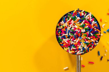 Yuvarlak metal bir kepçenin yukarıdan aşağı görüntüsü. Temiz sarı bir zemin üzerine renkli gökkuşağı parçacıkları serpiştirilmiş. Editoryal gıda içeriği veya tarif çizimleri için harika.