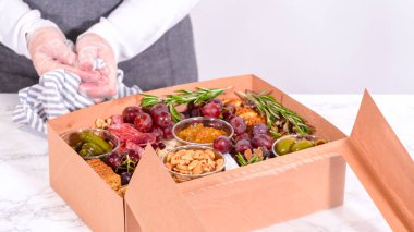 Elleri titizlikle bir kaseye taze kırmızı üzüm ekliyor, çeşitli peynirler, zeytinler ve pişirilmiş etlerle dolu güzel bir şarküteri kutusunu tamamlıyor.