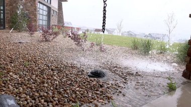 Yağmur suyu, manzaralı bir banliyö arka bahçesindeki yağmur zincirinin içinden akar. Su, çakıl yatağına ulaştığında görüntü sıçrar ve hareket eder..