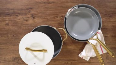 Kırsal ahşap bir yüzeye sergilenen zarif seramik tencere seti, beyaz kapaklı bir tencere ve göz alıcı altın saplı iki gri çubuksuz tava içeriyor. Beyaz ve kırmızı çizgili bir mutfak havlusu...