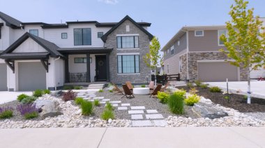 Yeni inşa edilen banliyö evlerinde dekoratif taş yolları, çalıları ve küçük ağaçları olan manzaralı ön bahçeler yer alıyor. Evler temiz bir mimari tasarıma sahip ve parlak güneşli bir havada fotoğraflanıyorlar.